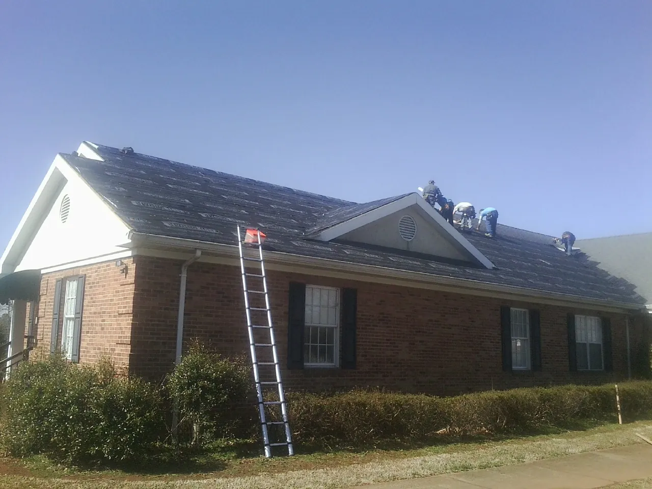 Crew tearing shingles off a brick ranch roof with a ladder set — Metal Roof Repair work by our crew in Skillman