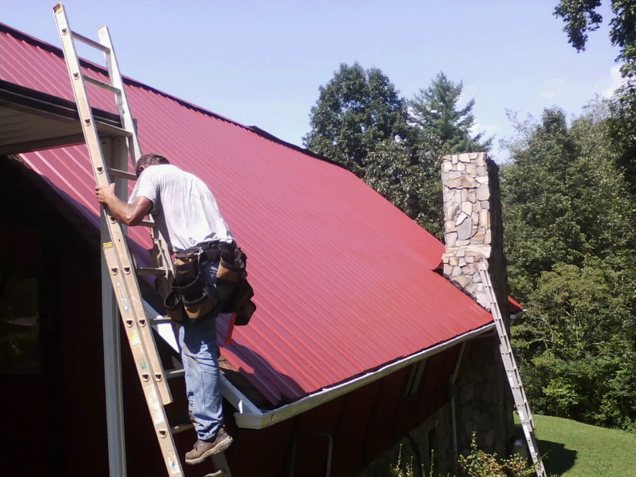 Roofer on a ladder beside a red metal roof and stone chimney — Soffit & Fascia Repair by Roofing Brothers in Skillman